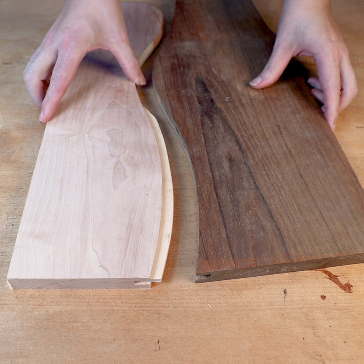 Hands holding two curved wooden boards, light and dark hardwood, on a workbench.