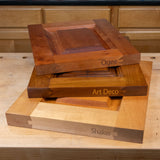 Three stacked wooden cabinet door panels labeled Ogee, Art Deco, and Shaker on a workbench.