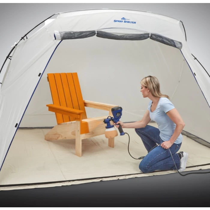 Woman using paint sprayer on wooden chair in portable spray shelter for woodworking finishing