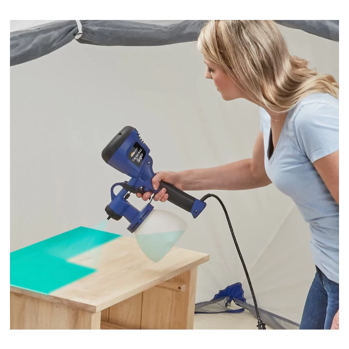 Woman using electric paint sprayer to apply finish on wooden table in workshop