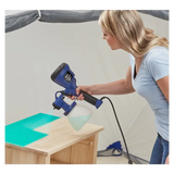Woman using electric paint sprayer to apply finish on wooden table in workshop