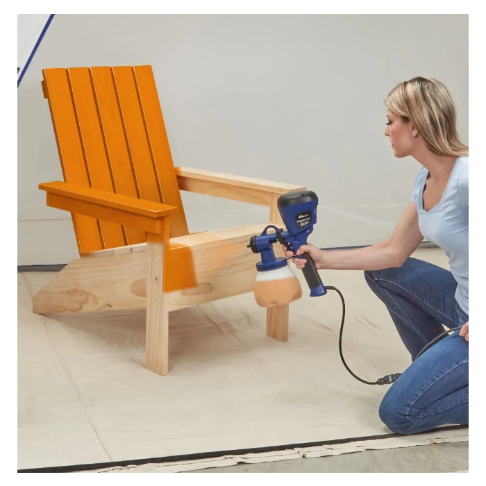 Woman using paint sprayer to finish wooden Adirondack chair in workshop setting