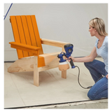 Woman using paint sprayer to finish wooden Adirondack chair in workshop setting