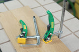 Two green toggle clamps securing a wooden board on a workbench, woodworking tools