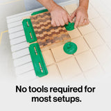 Hands securing a wooden board with green CNC hold-down clamps on a grid table.