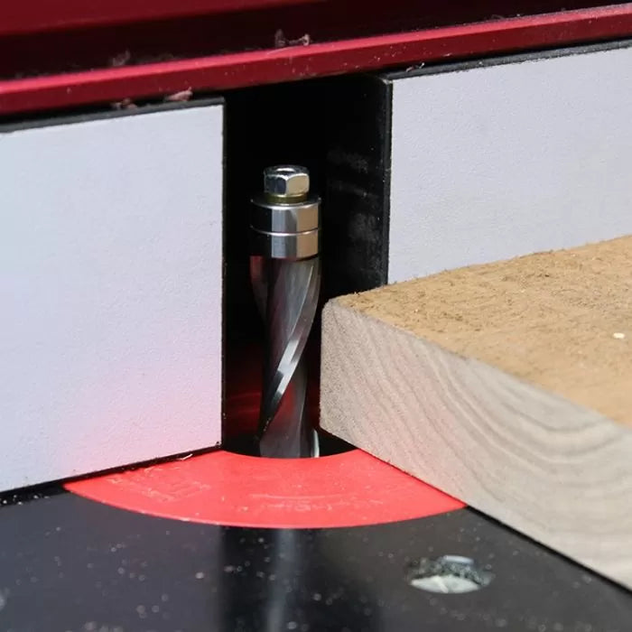 Spiral router bit cutting wood on a router table with red insert ring and fence.