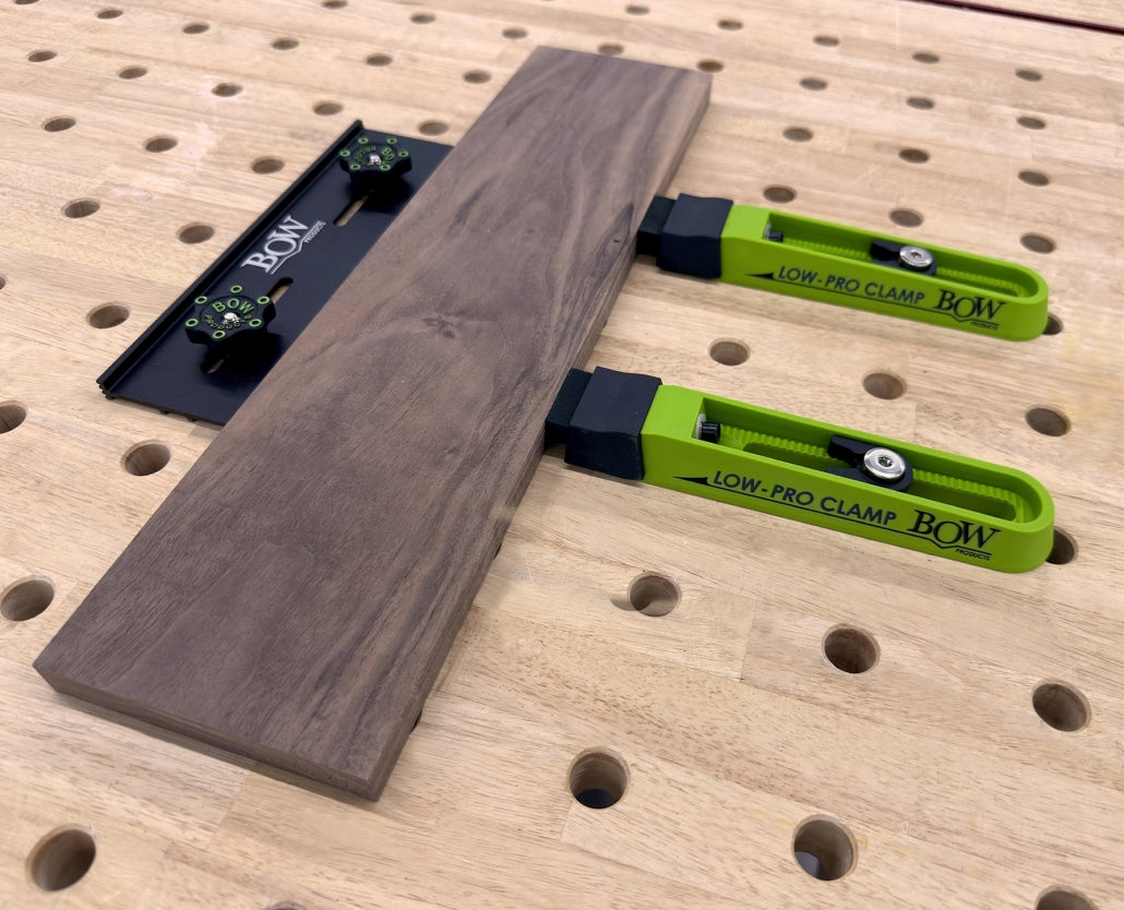 Two green low-profile woodworking clamps securing a board on a perforated workbench.