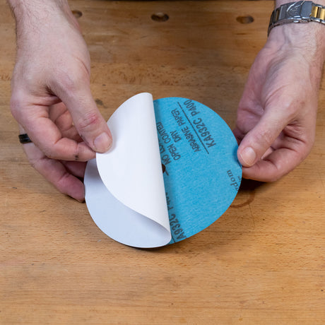 A close-up of a person's hands peeling back the white backing paper from a circular blue sanding disc on a wooden workbench.