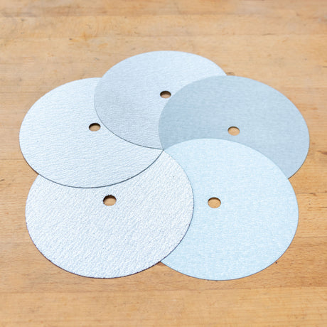 Five circular sanding discs with holes, arranged in a cluster on a light brown wooden surface.