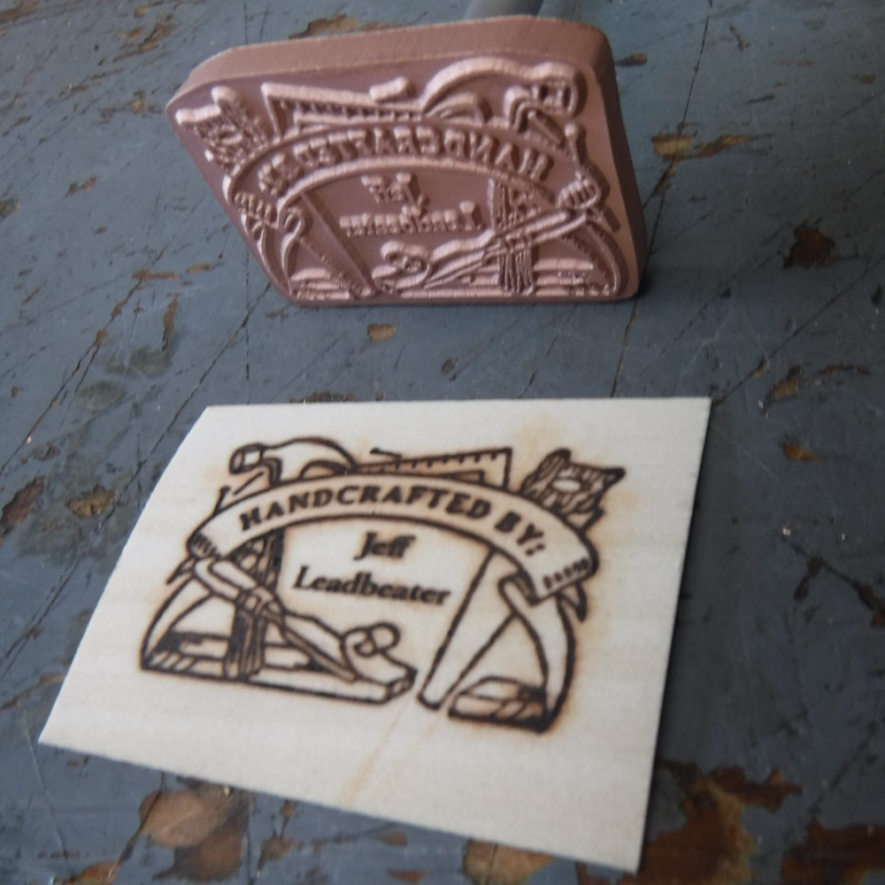 Woodworking branding iron and wood stamp reading 'Handcrafted by: Jeff Leadbeater' on workbench