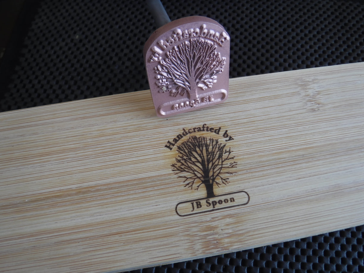 Custom branding iron with tree design marking 'Handcrafted by JB Spoon' on wood.