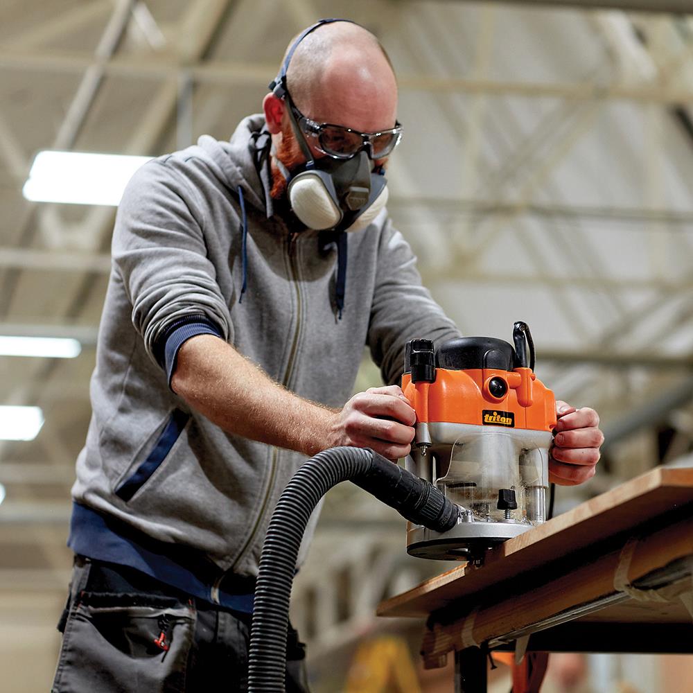 Woodworker using an orange router tool on wood, wearing safety goggles and dust mask