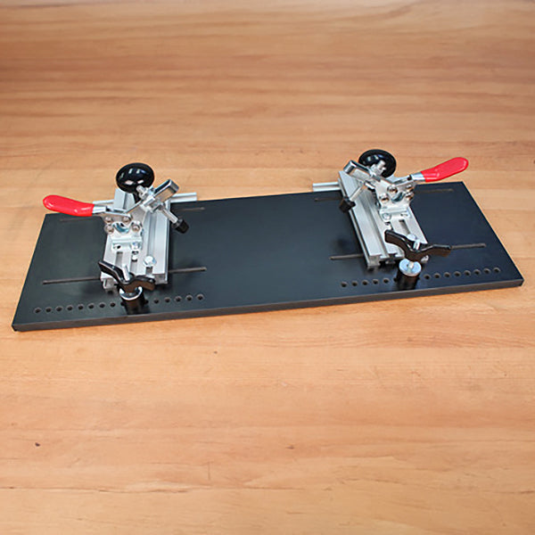 Woodworking jig with two adjustable clamps on a black metal plate, set on a wooden workbench