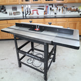Woodworking router table with fence in a workshop, surrounded by tools and cabinets.
