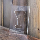 Transparent acrylic template with martini glass cutout and engraved text, on wooden background