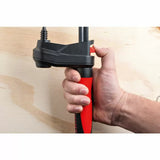 Hand gripping a woodworking clamp with a red and black ergonomic handle on plywood background