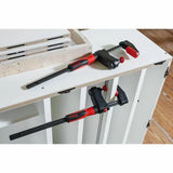 Two bar clamps holding white cabinet parts on a woodworking workbench