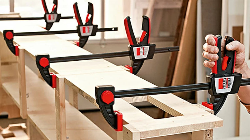Woodworking bar clamps securing boards in a workshop, close-up of clamp handles and frames.
