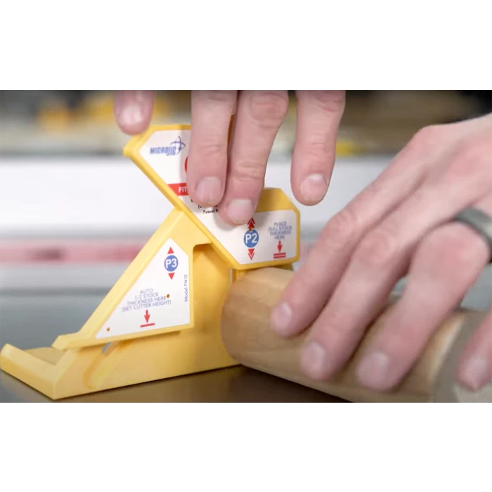Hand using a yellow woodworking gauge tool to measure a round wooden dowel on a workbench.