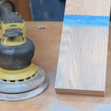 Orbital sander next to sanded wood board with blue painter’s tape, woodworking tools