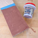 Brown wood grain filler applied to a wooden board, container of filler beside it on workbench