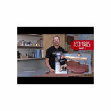 Woodworker in workshop presenting live-edge slab table project, woodworking tools in background.