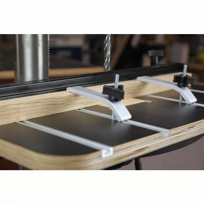 Woodworking drill press table with T-track hold-down clamps and drill bit in workshop
