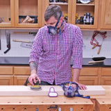 Woodworker sanding board with dust mask in workshop, surrounded by woodworking tools
