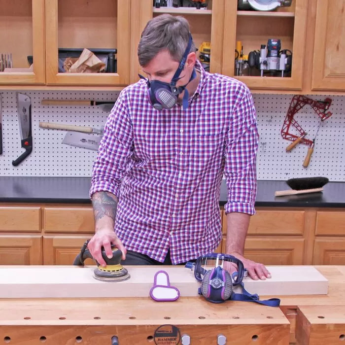 Man sanding wood with a power sander in a woodworking shop, wearing a dust mask for safety.