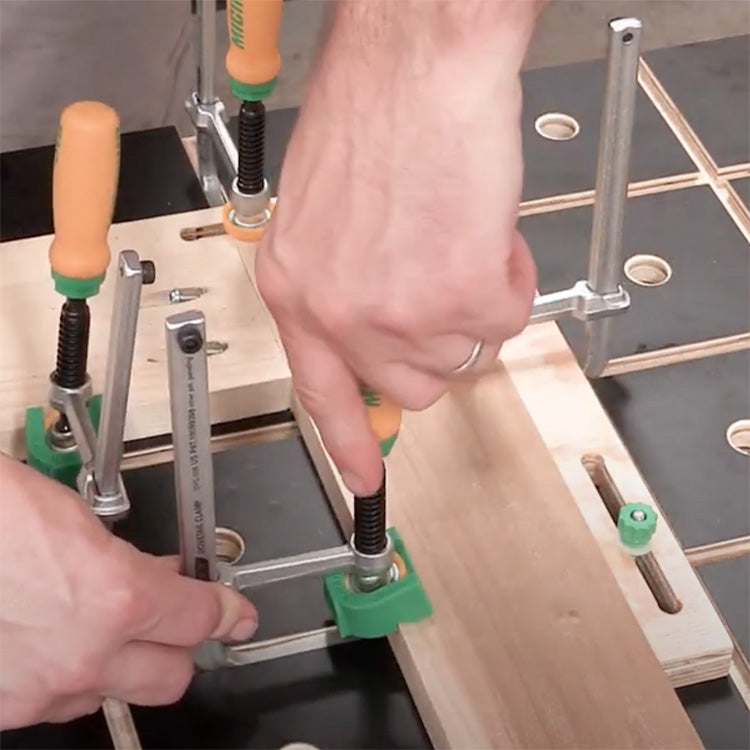 Woodworker using F-style clamps to secure wood on a perforated workbench for precision woodworking.
