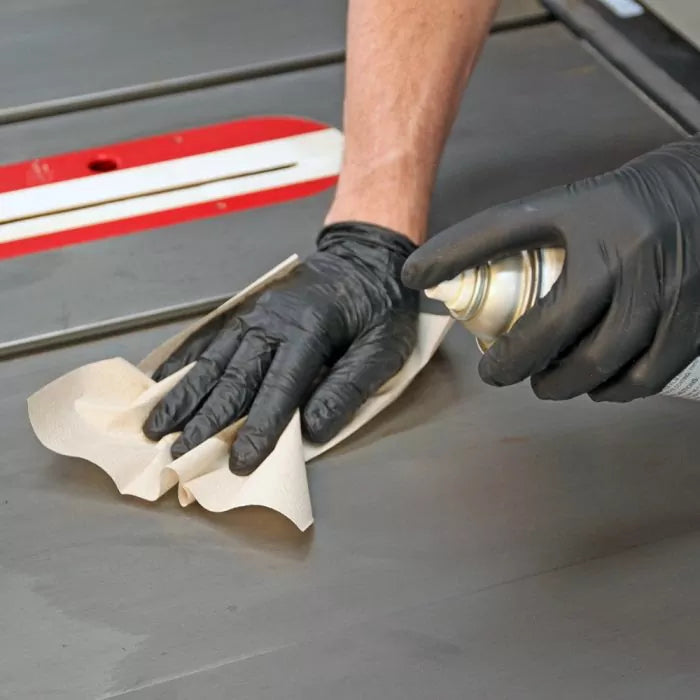 Person wearing black nitrile gloves applies spray lubricant to a table saw surface with a cloth