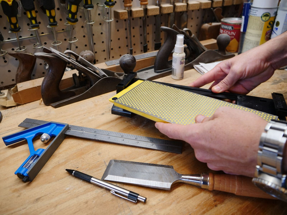 Woodworker sharpening chisel with diamond sharpening stone on a workbench, surrounded by hand tools