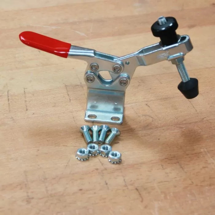 Metal toggle clamp with red handle and mounting hardware on a wooden workbench